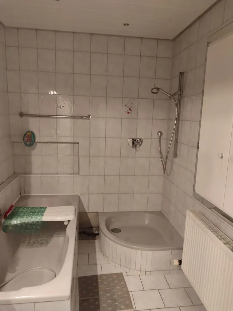 Altes Bad mit weiß gefliesten Wänden, Badewanne und flacher Eckduschwanne vor dem Umbau
