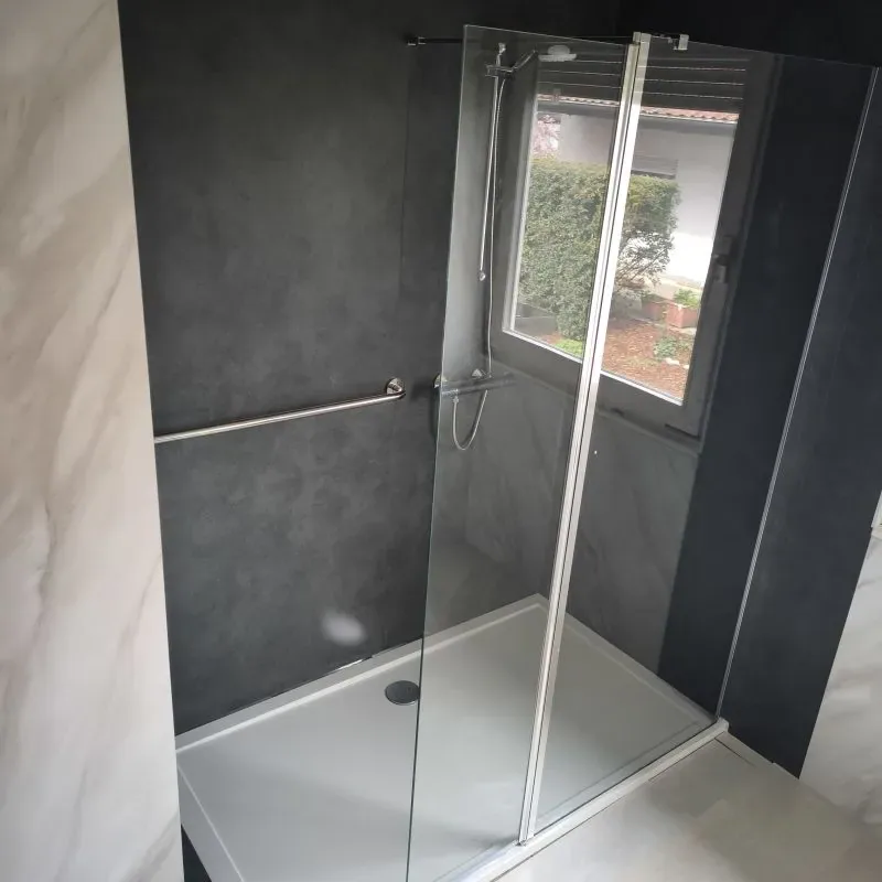 Bodengleiche Dusche mit anthrazitfarbenen Wandflächen, Haltegriff und rahmenloser Glasscheibe vor einem Fenster
