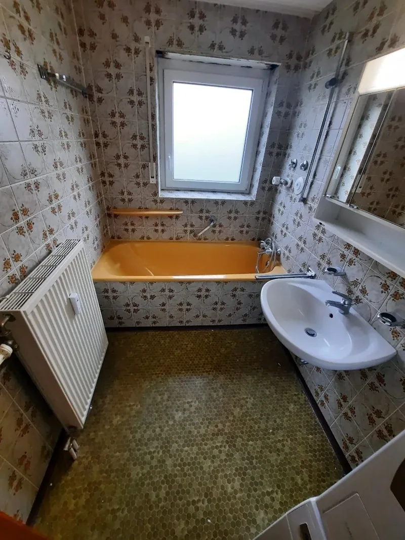 Altes Bad mit oranger Badewanne, gemusterten Fliesen und grünem Bodenbelag vor dem Umbau